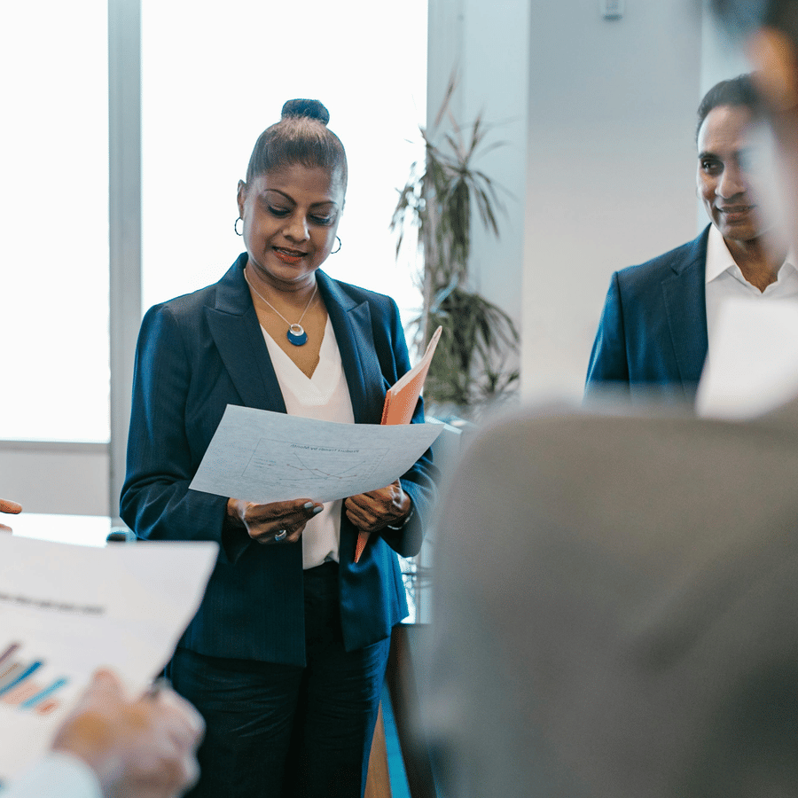 Professionally dressed individuals in a modern office meeting, discussing documents with colorful bar graphs, as one person presents while others review papers in natural light.