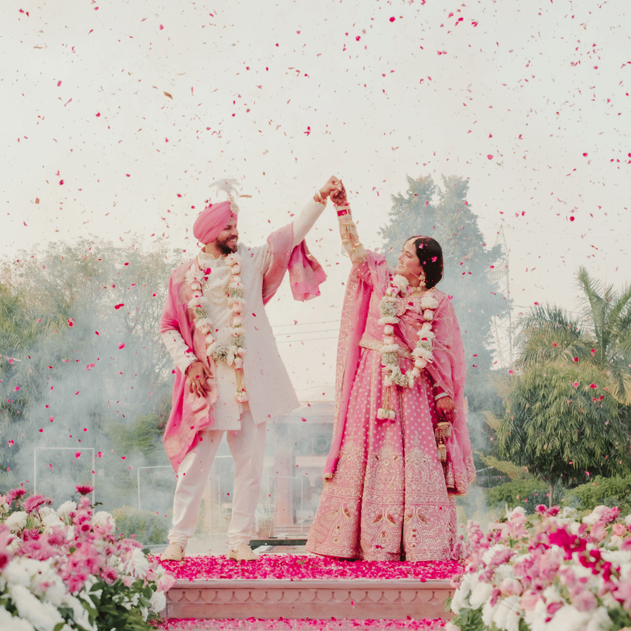 A newlywed couple holds hands and raises their arms on a stage decorated with white and pink flowers during an outdoor wedding ceremony at Umaid Palace.