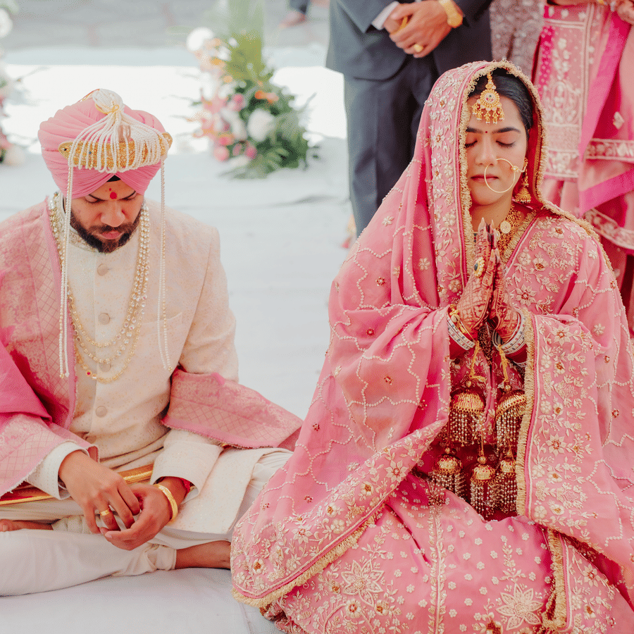 A bride with hands folded in prayer sits next to her groom during their traditional wedding ceremony at Umaid Palace.