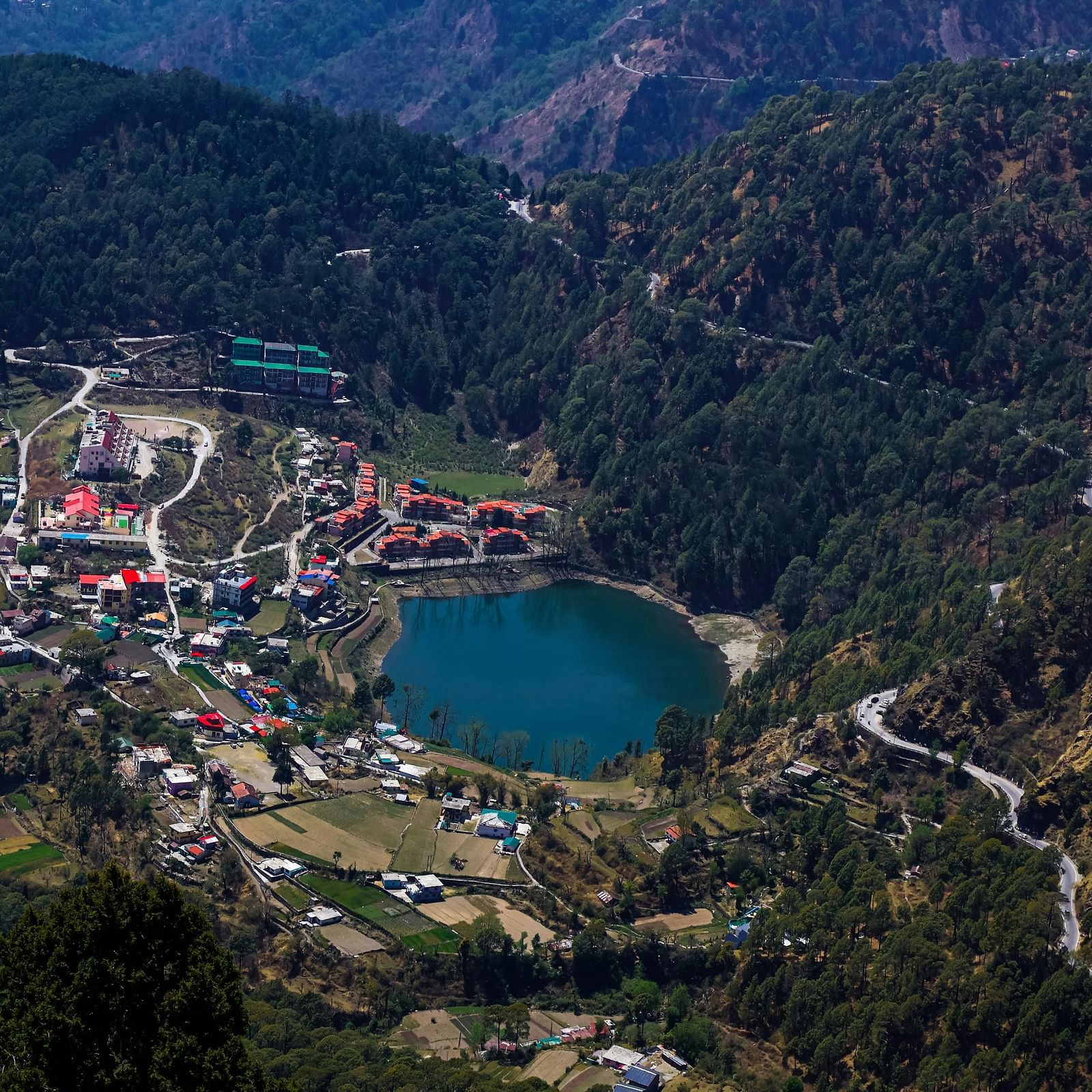 A scenic view of Nainital city  with a lake surrounded by huts and lush green hills in the background.