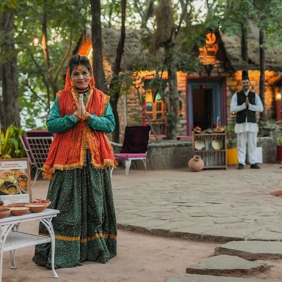 A person wearing a colorful outfit standing in a clearing with buildings and trees in the background.