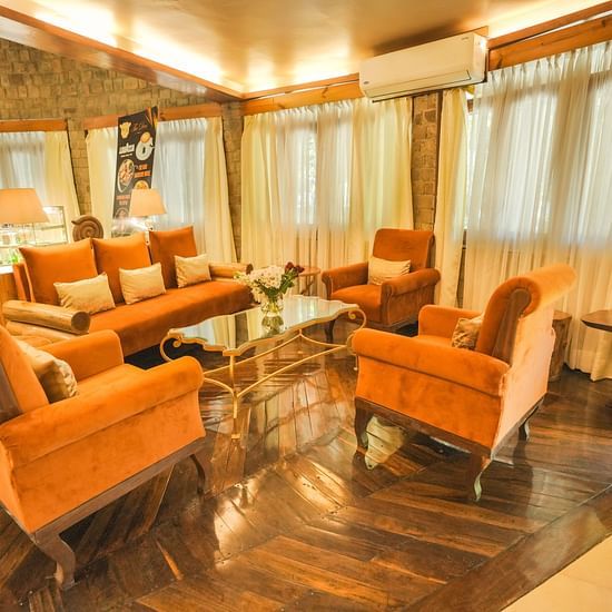 A room with orange armchairs, a couch, and large windows, warm lighting.