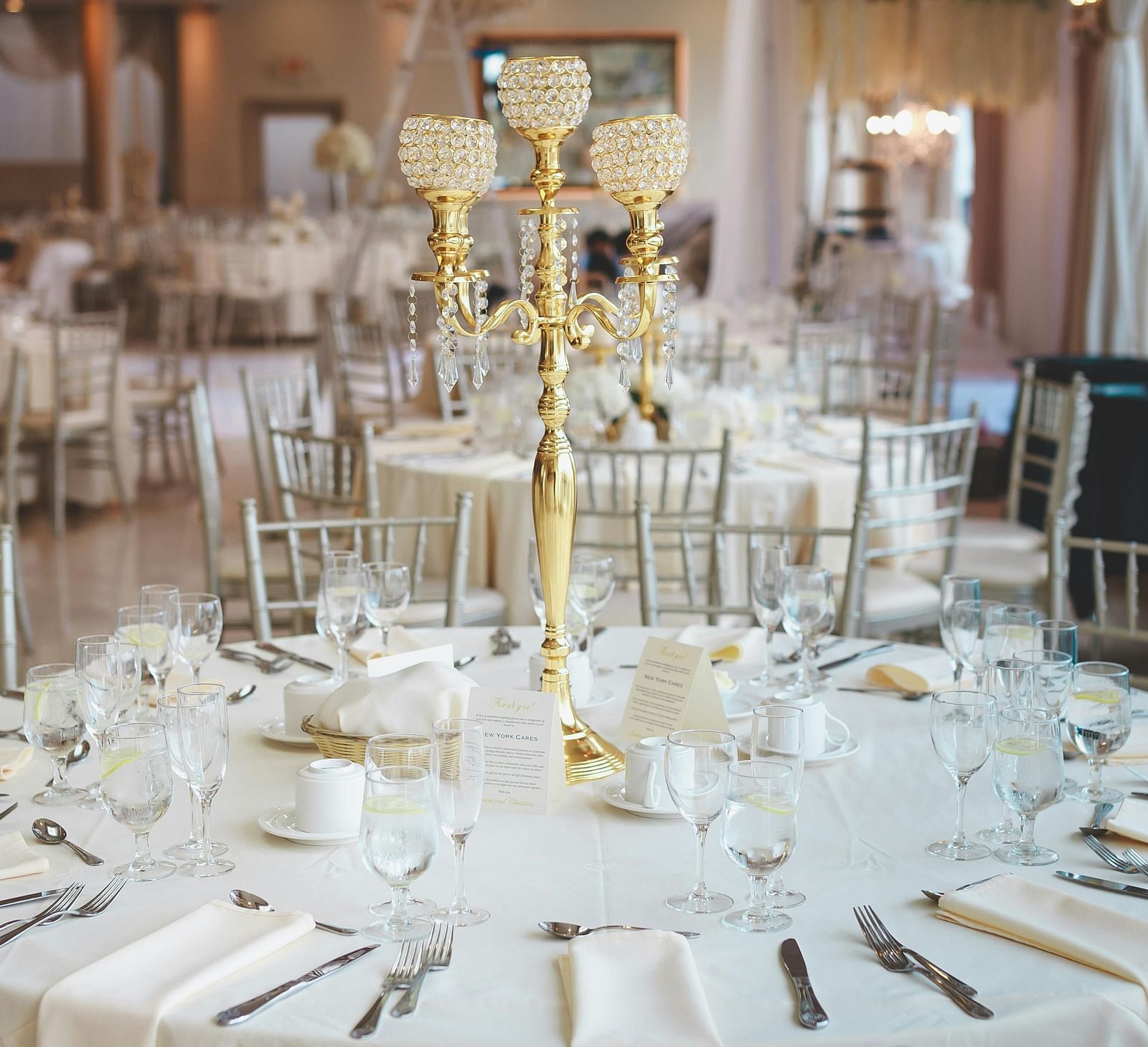 A formally set round table, featuring a white tablecloth, elegant place settings with silverware, glassware, and napkins, and a tall, gold candelabra as a centrepiece, all set within a large room with additional tables and chairs in the background.