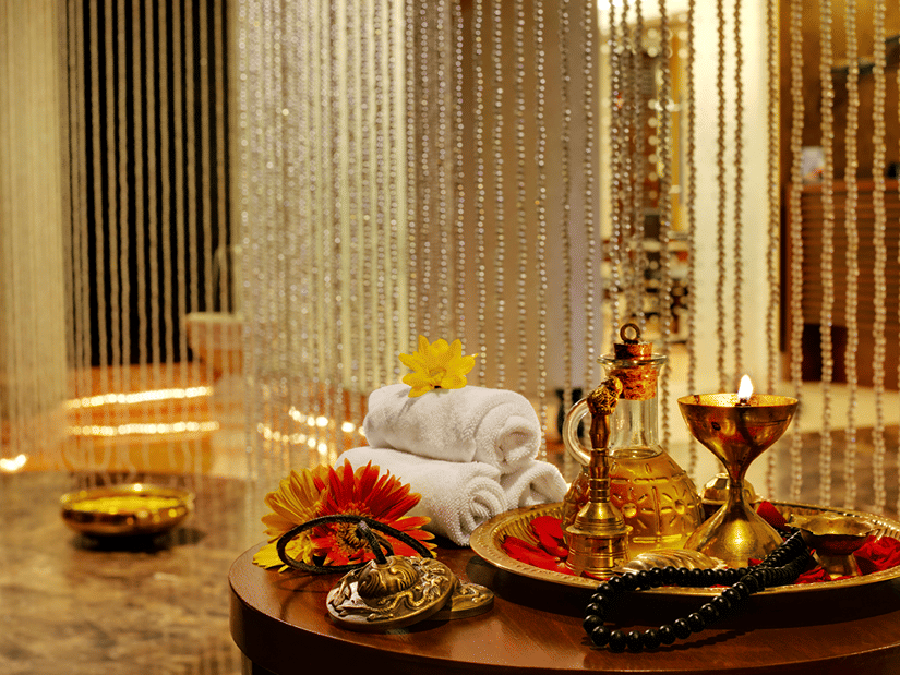 Myra Spa at Kenilworth Resort & Spa, Goa, featuring rolled white towels, a brass tray with massage oil, a lit lamp, fresh flowers on a round wooden table, and shimmering beaded curtains in the background.