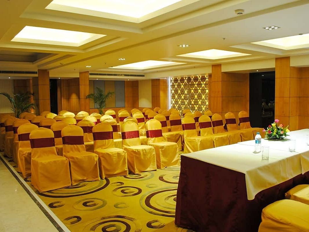The image shows a conference room with rows of chairs facing a table with water bottles and flowers - Lords Eco Inn Navsari.