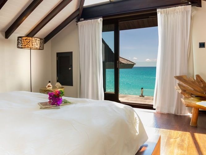 Cozy beachfront bedroom with vaulted ceilings, white linens, and open curtains revealing stunning ocean views just steps from the private terrace.