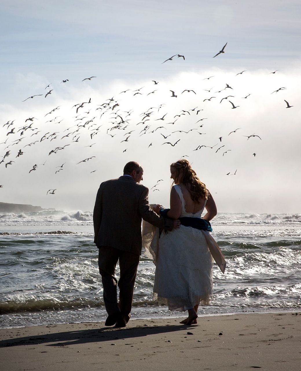 Newlyweds strolling on the beach as seagulls take flight around them