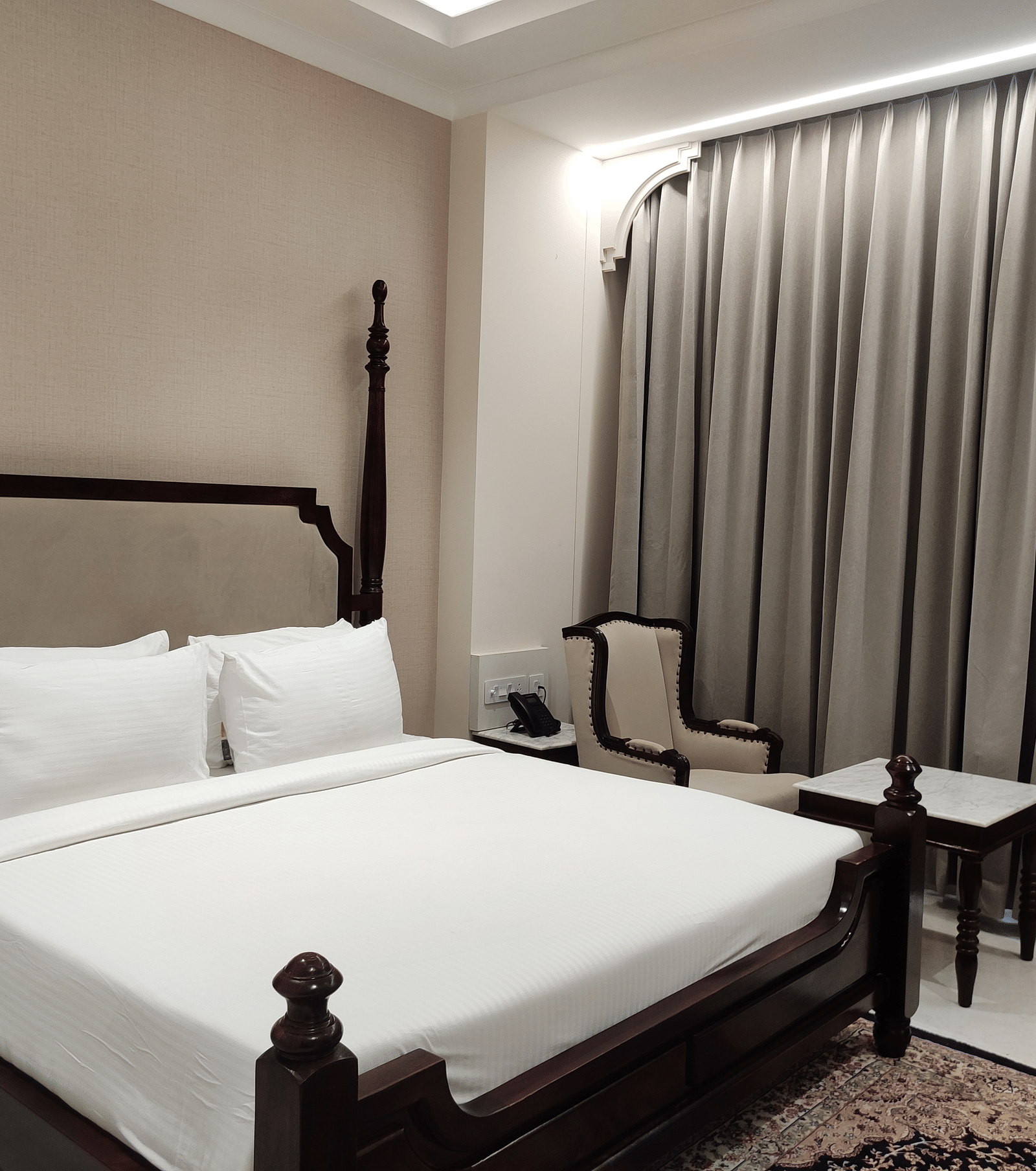 A bedroom at Anant Mahal, Jaipur featuring a king size bed with multiple pillows arranged at the head of the bed, a patterned rug, floor to ceiling curtains behind a table, and a chair.