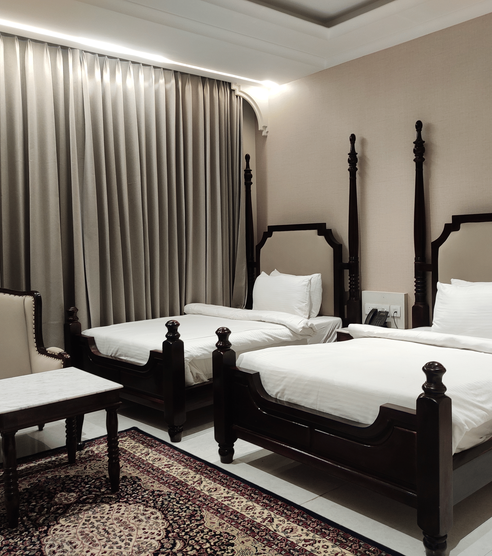 A bedroom at Anant Mahal, Jaipur featuring 2 double beds, a patterned rug, and long curtains covering a large window behind the seating area.