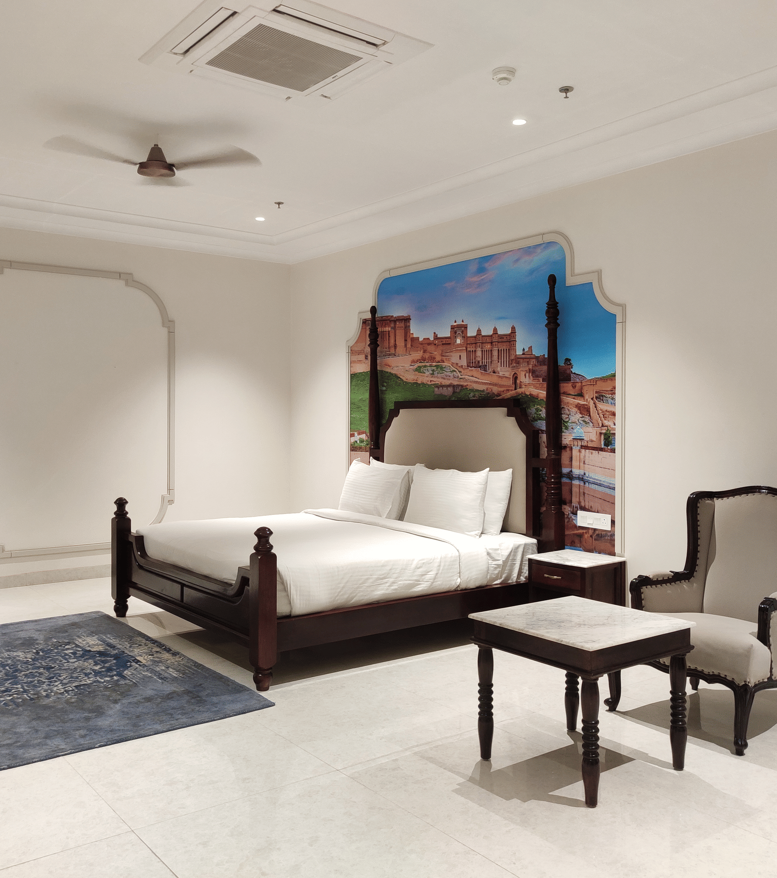 A bedroom view at Anant Mahal, Jaipur featuring a king size bed, a blue rug, a mirror, a seating area with a small table and a cushioned chair.