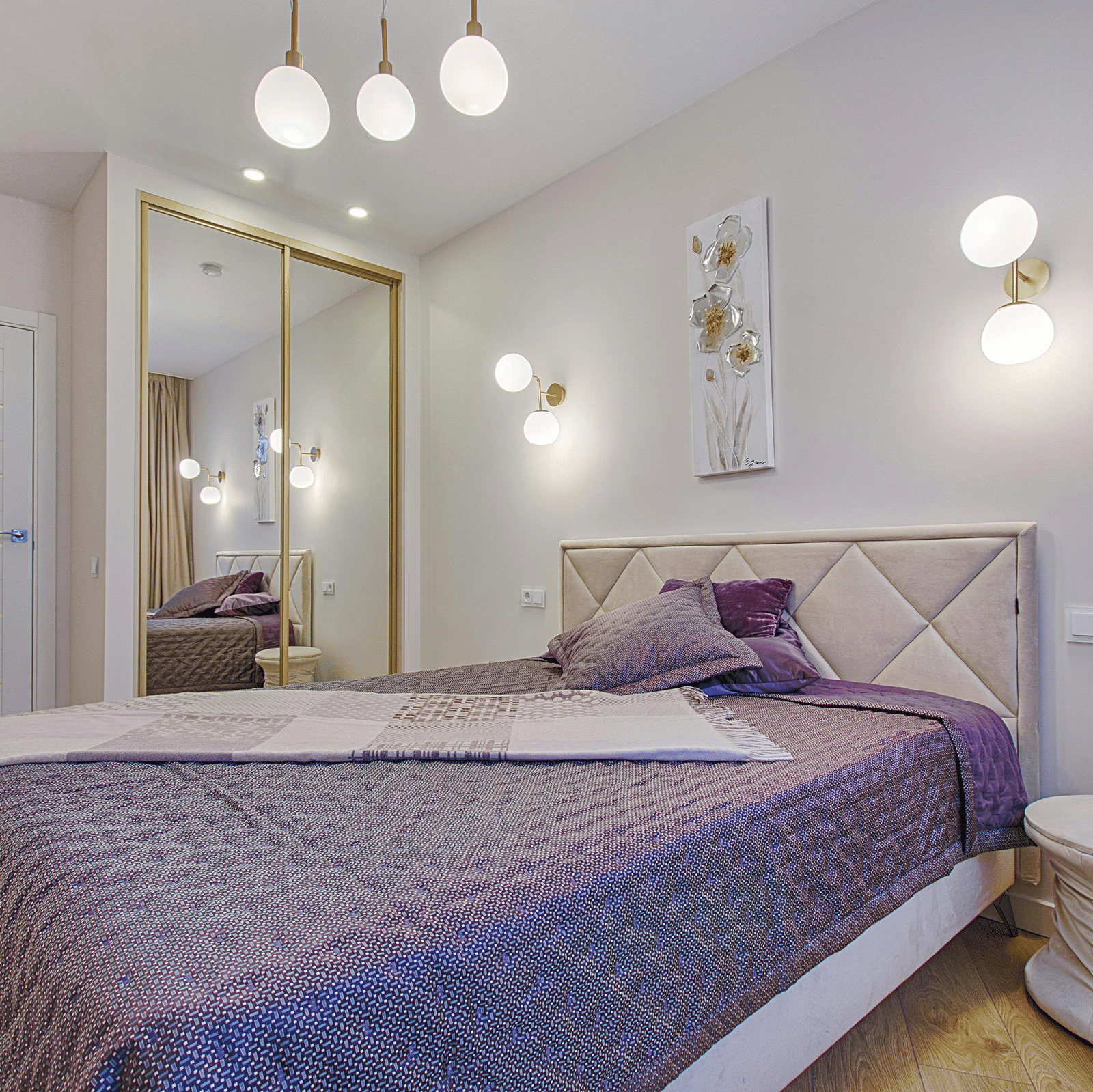 A minimalist bedroom with a quilted purple bedspread, a diamond tufted headboard, and mirrored wardrobe doors.