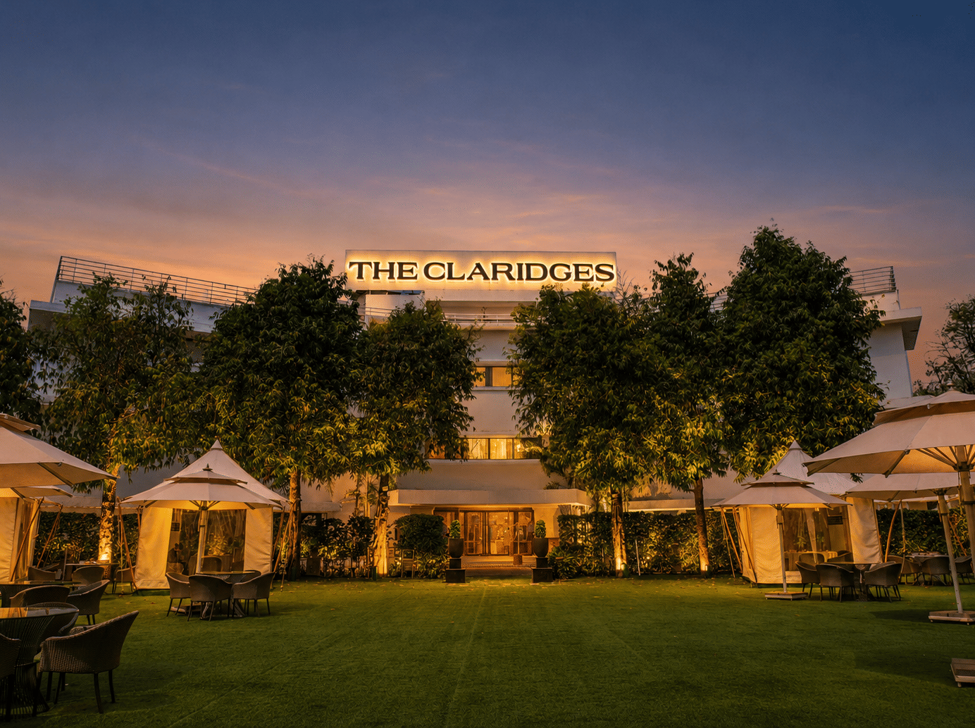 Evening view of a green hotel lawn featuring warm-lit white canopies and outdoor dining tables in front of an illuminated building at dusk - The Claridges New Delhi