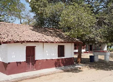 A small, simple building with a thatched roof sits amidst trees, casting shadows on the sandy ground.