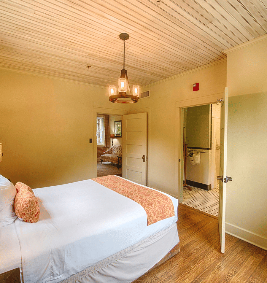 A wide view of a guest room with a bed, open doorway to bathroom, and a chandelier hanging from a wood-paneled ceiling.