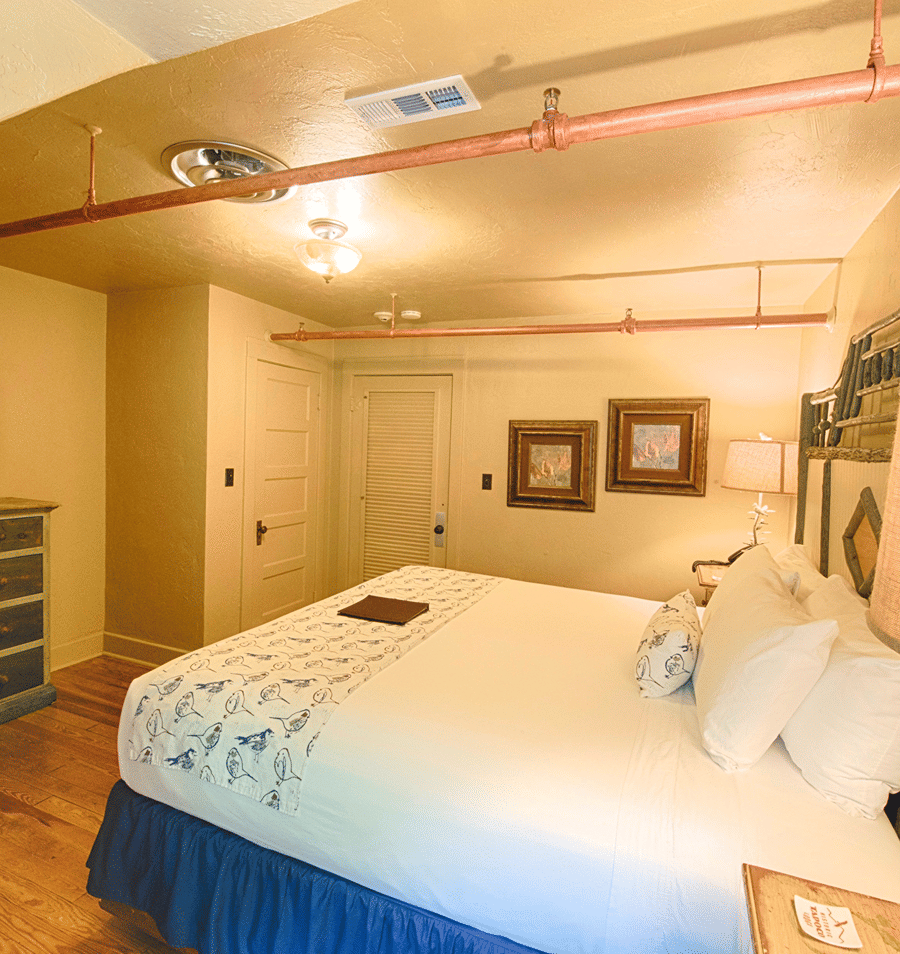 A guest room with a large bed, wooden floor, copper pipes, and artwork on the walls.