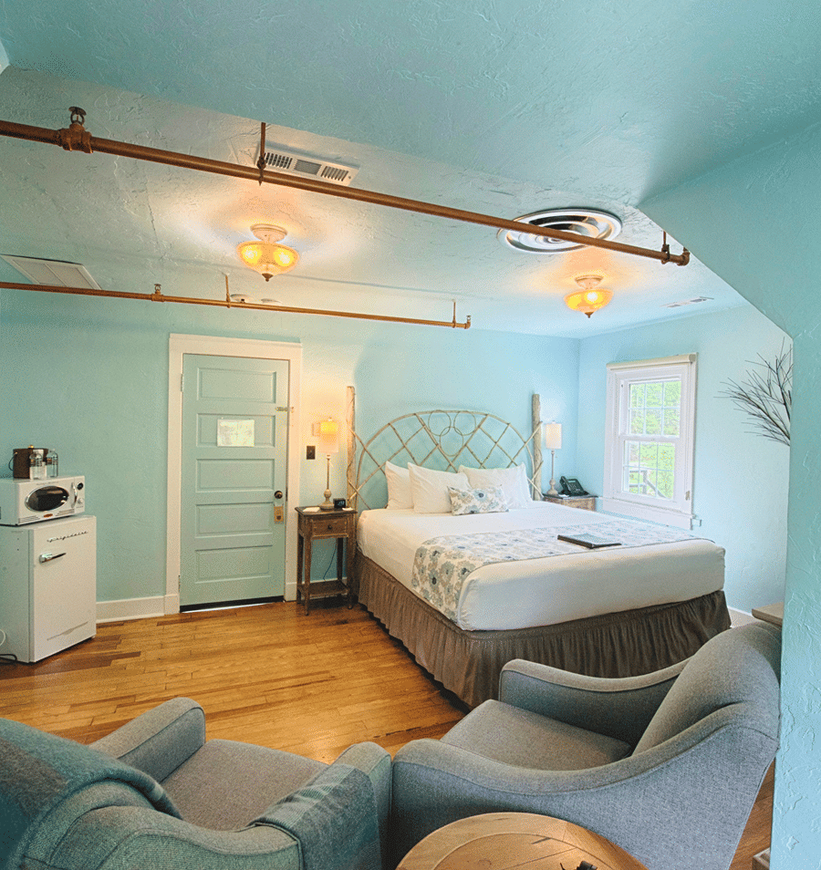 A wide view of a guest room with light blue walls, a king-sized bed, two armchairs, a mini-fridge, and a window.