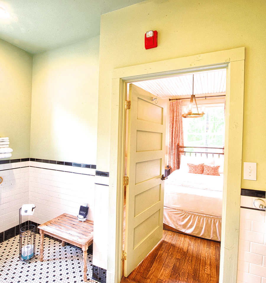 A bathroom with a tiled floor and walls, a wooden bench, and an open doorway leading to a bedroom with a bed.