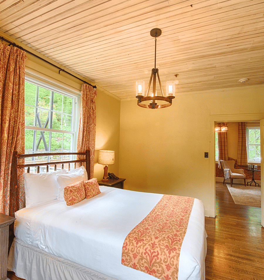 A neatly made bed with white linens and orange accents in a room with wood flooring and a chandelier.