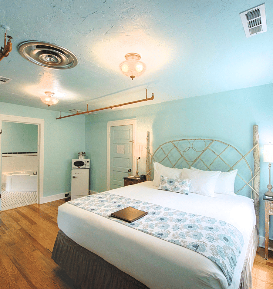 A wide-angle view of a hotel room with a large bed, nightstand, and mini-fridge.