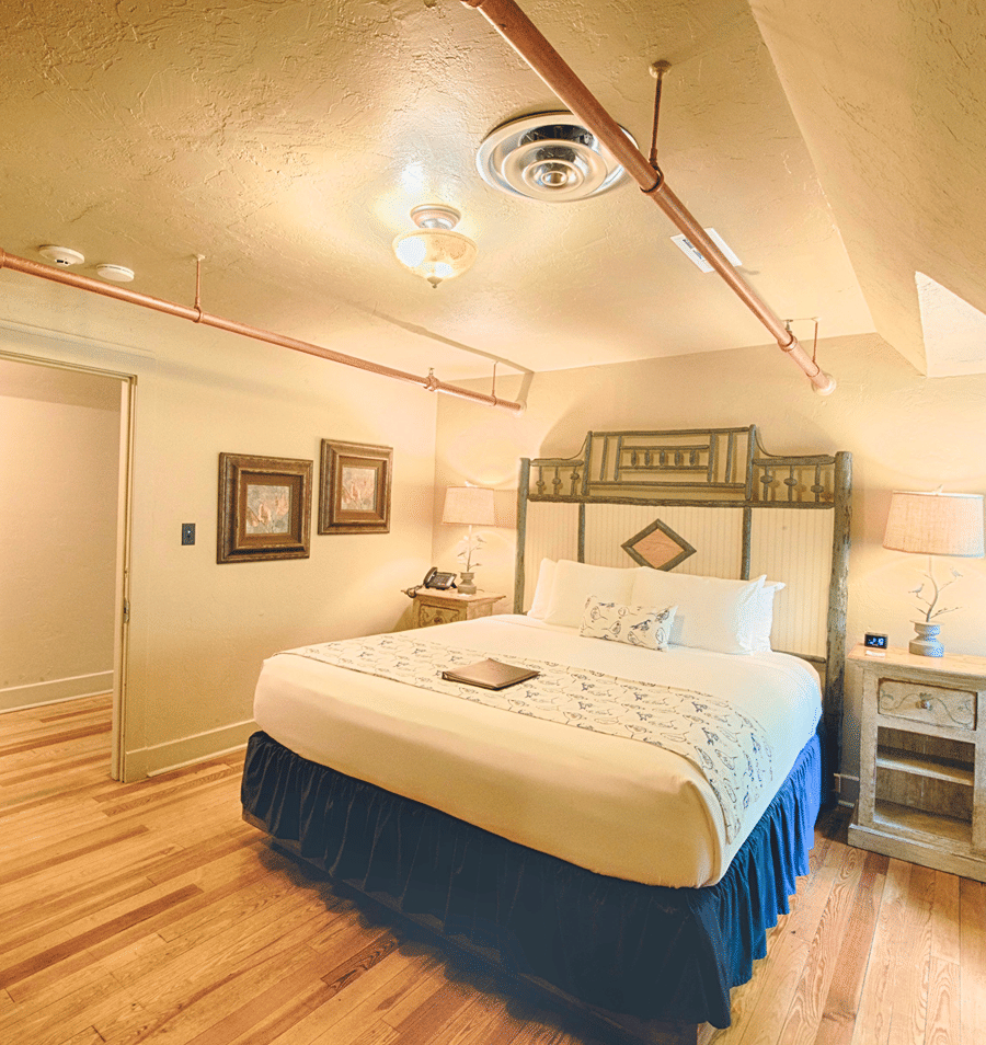 A hotel room with a large bed, wooden floor, and high ceiling with exposed pipes.