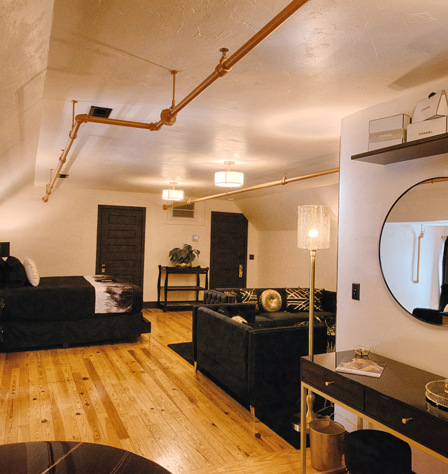 A wide view of a living room with a sofa, bed, desk, and exposed pipes on the ceiling.