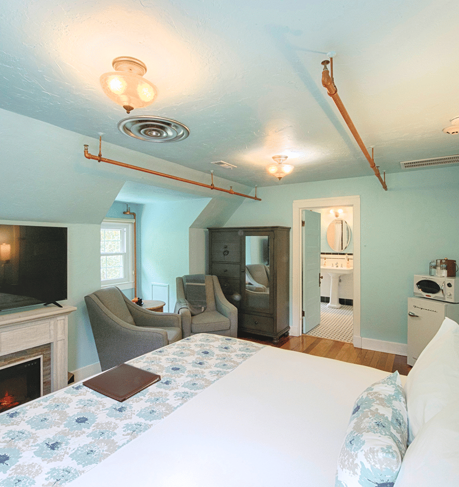 A wide view of a guest room with a bed, TV, fireplace, armchairs, and open bathroom door.