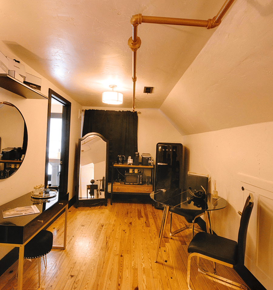 A wide-angle view of a guest room with a glass table, two black chairs, a mini fridge, and a round mirror on the wall.