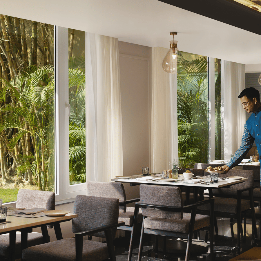 A waiter in traditional blue attire setting a table in a bright restaurant with floor-to-ceiling windows and garden views.
