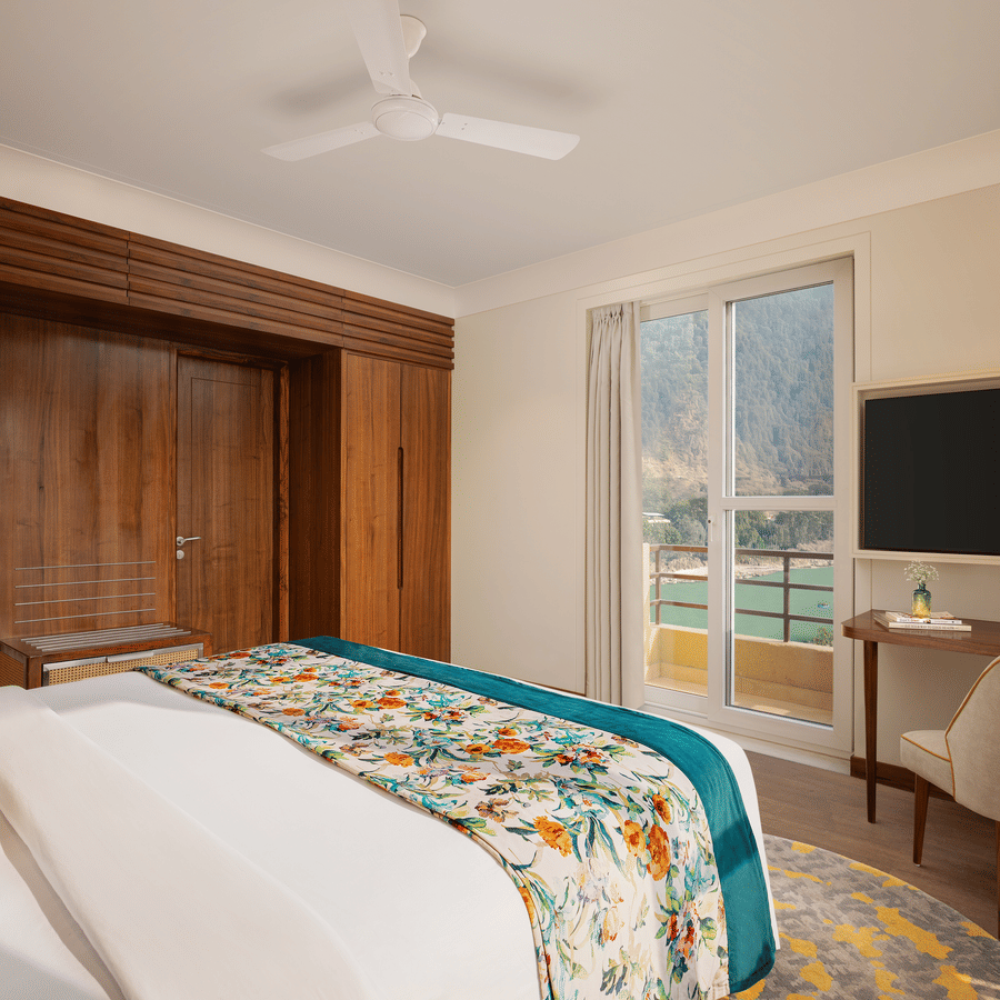 Bright hotel bedroom featuring floral linens, wooden paneling, a desk area, and a balcony with a scenic river view.