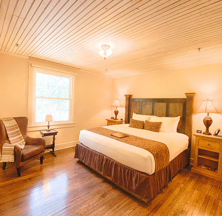 full view image of a brightly lit bedroom with a comfortable double bed with a heady headboard along with an armchair - Historic Tapoco Lodge