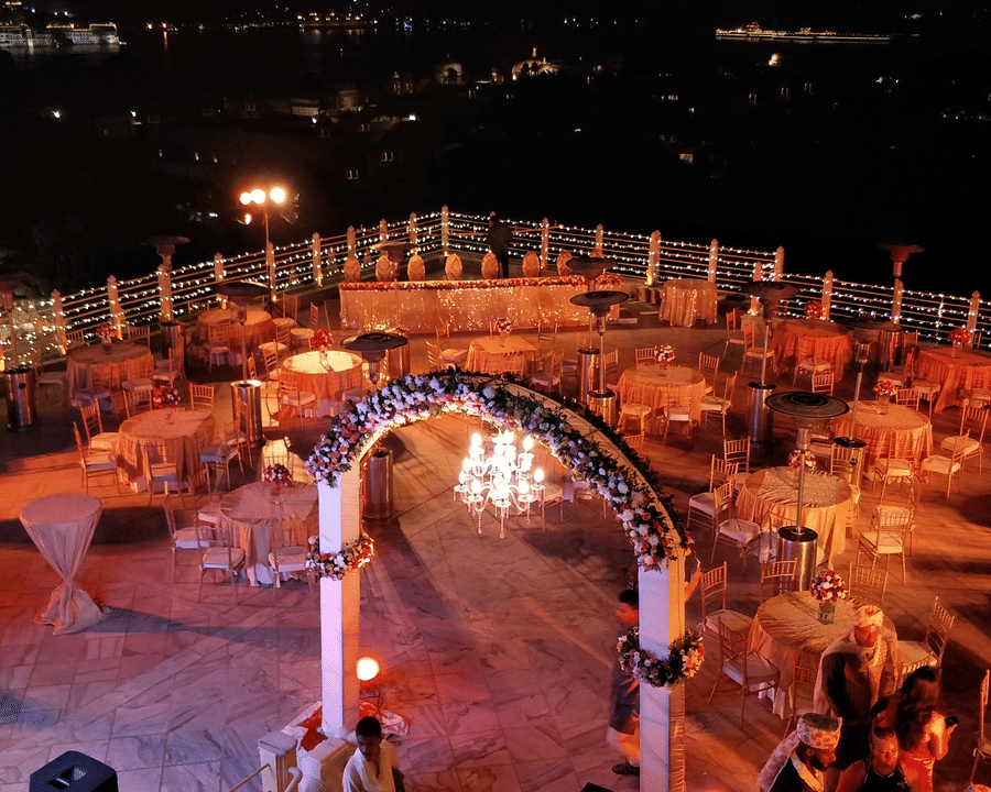 The open-air Square Terrace reception at Chunda Palace, Udaipur, beautifully lit with a floral entrance arch, a glittering chandelier, and dining tables.