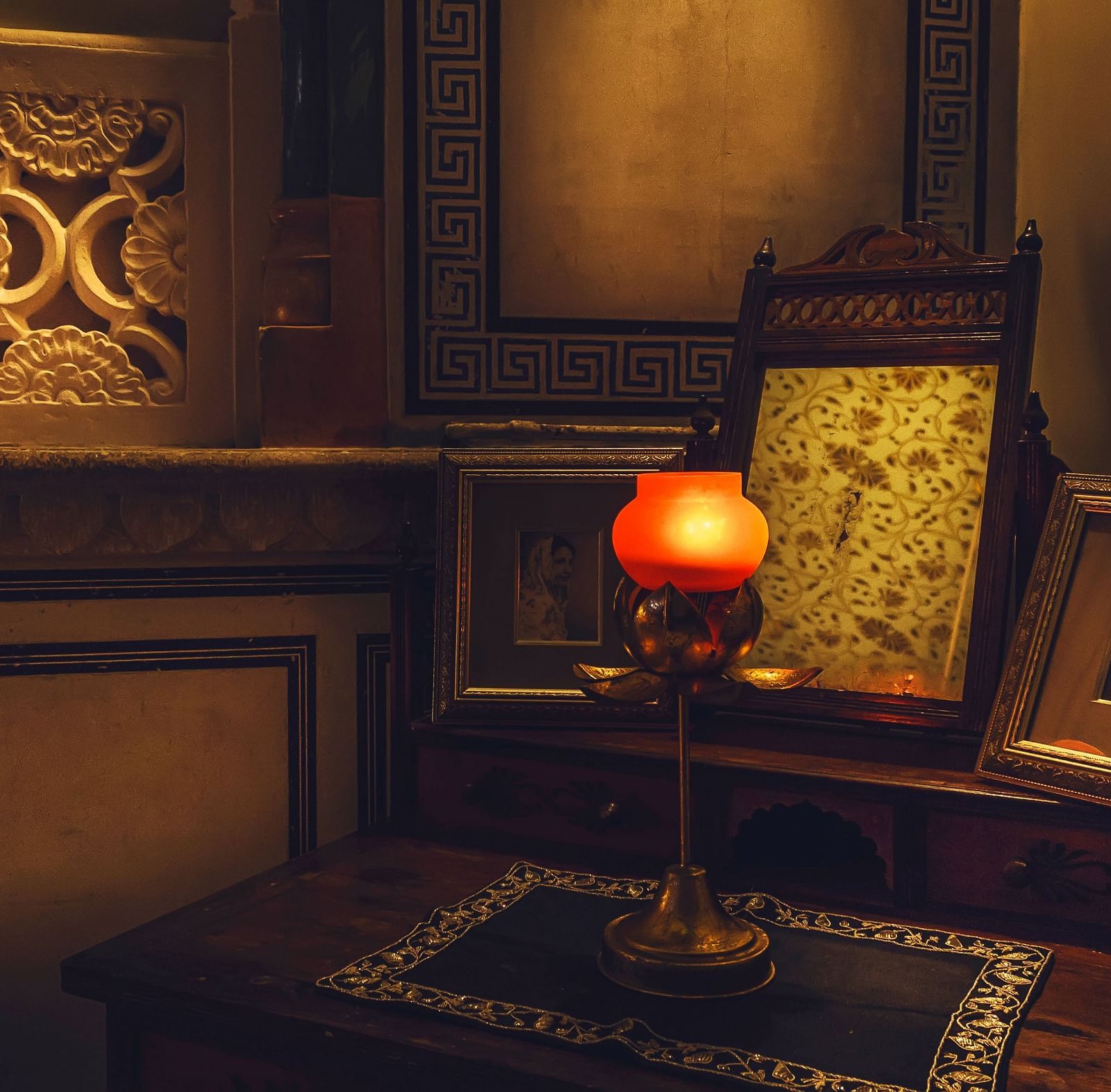 A dimly lit room at Umaid Vilas Jaipur featuring a decorative wall cutout, a lamp with an orange shade, and framed artwork.