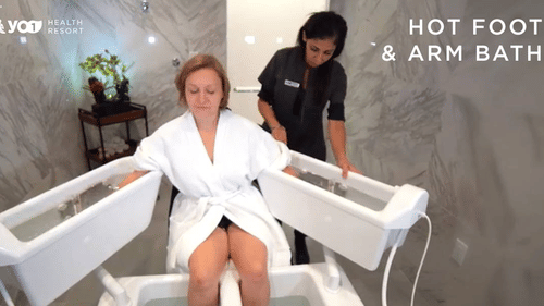 A woman in a white robe receives a hot foot and arm bath from a therapist in a modern spa setting at YO1 Longevity & Health Resorts