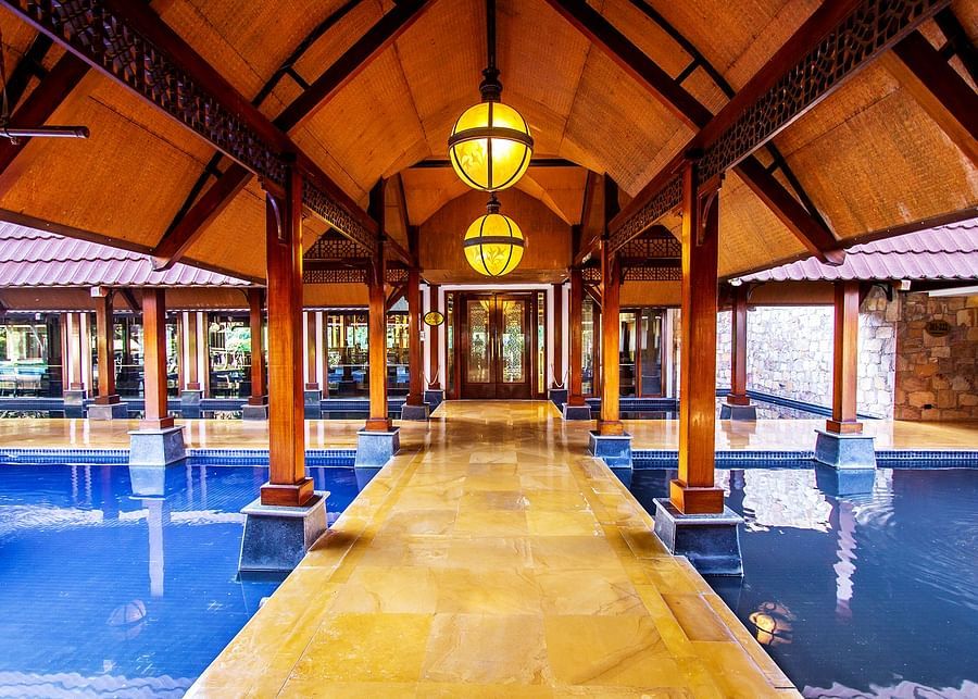 One of the interior walkways of Ananta Elite Spa & Resort bordered by a water body and with wood works 
