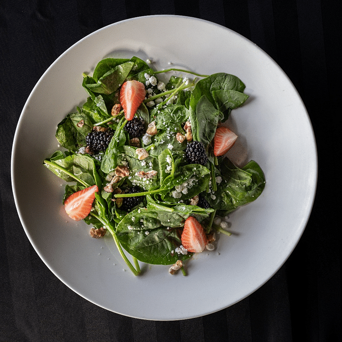 Fresh spinach salad served in a white bowl, garnished with strawberries, blackberries, nuts, and seeds on a dark background for a vibrant presentation