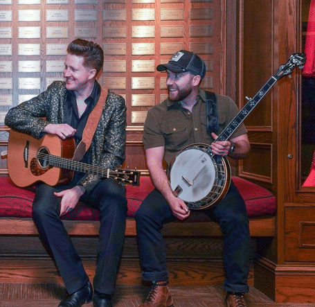Four men sit on a red bench with musical instruments: a violin, mandolin, guitar, and banjo. They are smiling warmly, indicating camaraderie.