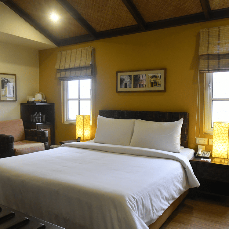 Deluxe Room at The Richmond, Puducherry featuring a plush bed with fresh linen and two lamps placed on the both sides on the nightstand as the sunlight makes its way through the two windows next to a sitting space