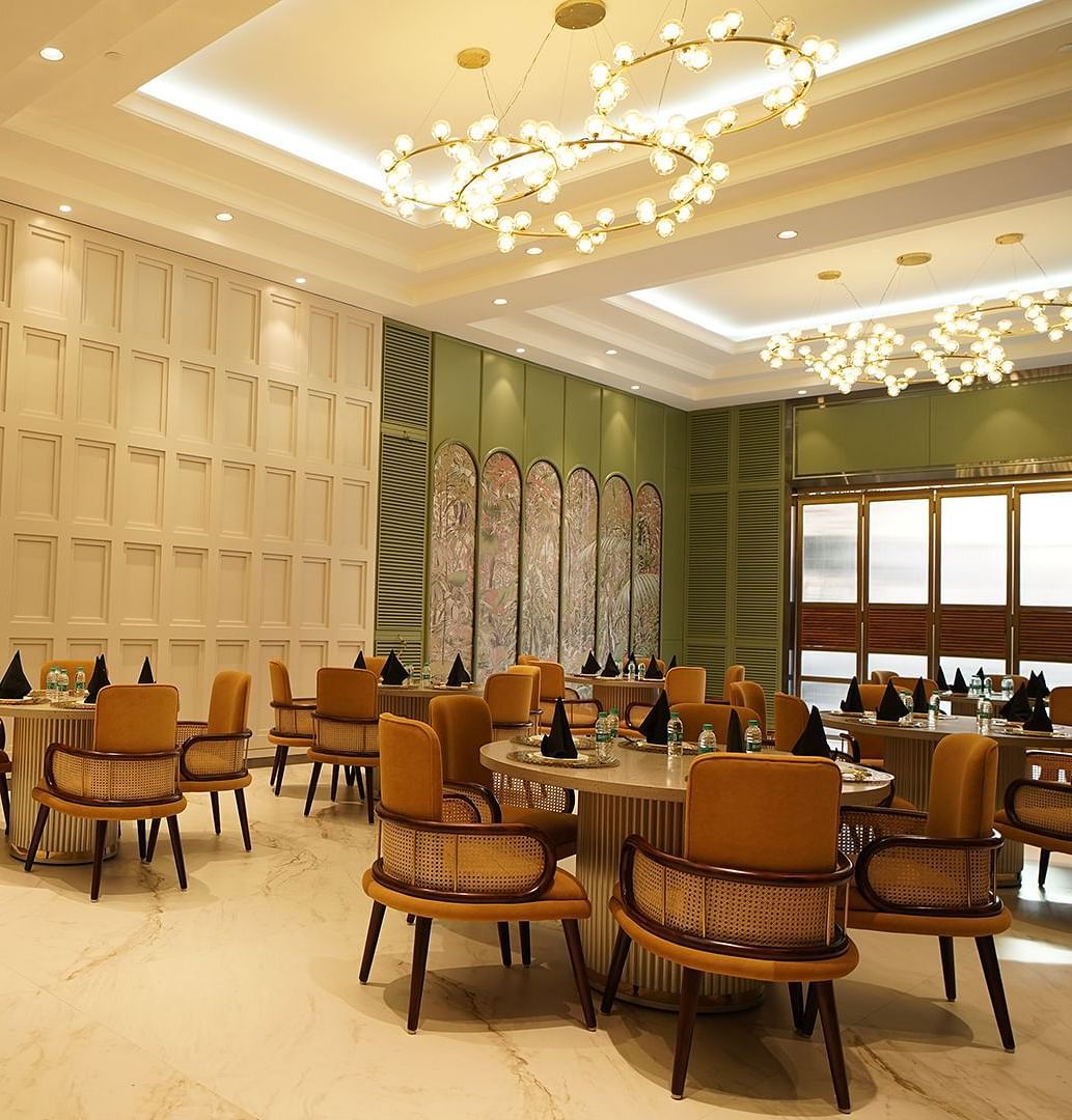 A stylish dining area at Nirvana Luxury Hotel, Ludhiana, featuring round tables with tan chairs, modern chandeliers, and elegant wall decor.
