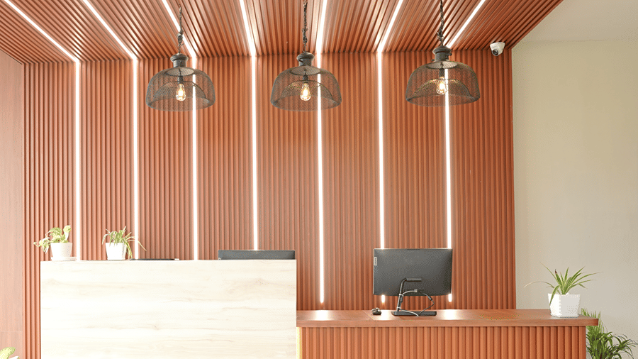 Close-up of the reception desk at Pride Elite Mandarmani with a wooden counter, monitor, 3 cage pendant lights, ribbed wood panelling, and potted plants.