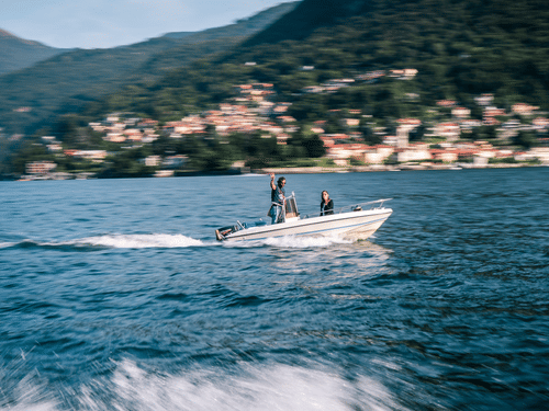 A small motorboat with 2 people on board moves across blue water, leaving a white wake, with a hillside town in the background