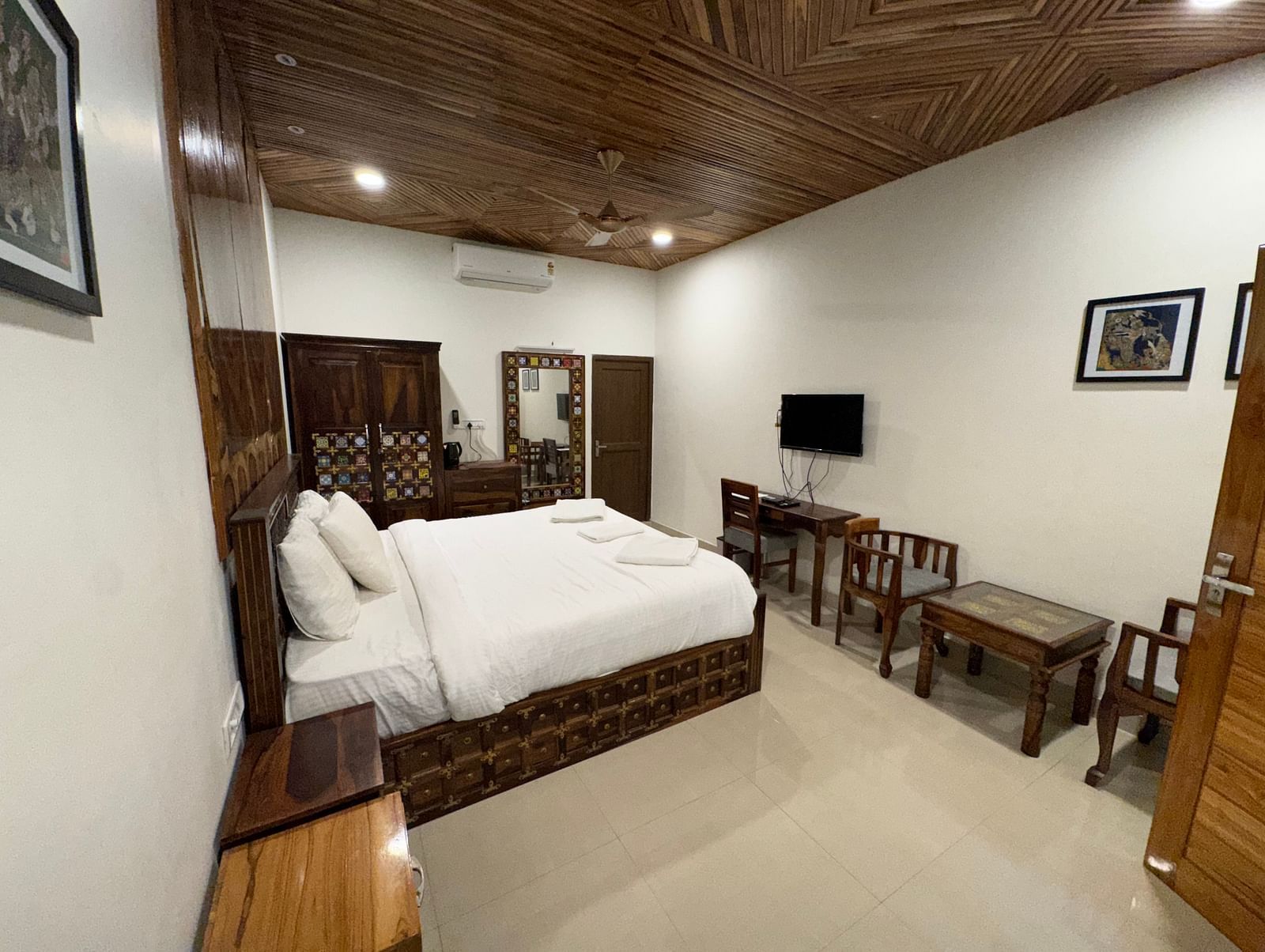 A hotel bedroom featuring a large white bed, dark wooden furniture including a TV cabinet, and a textured wooden ceiling.