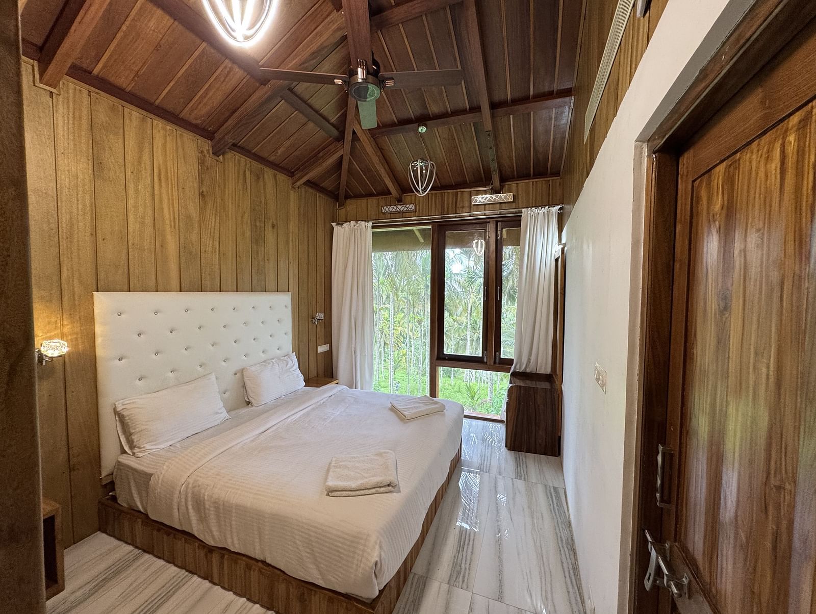 A resort bedroom with two beds, wooden walls and ceiling, a ceiling fan, and large windows with curtains.