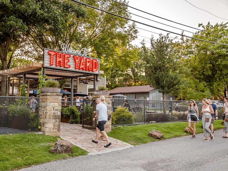 Man walking into a place with a board titled the yard while a group of women walk through a paved road.