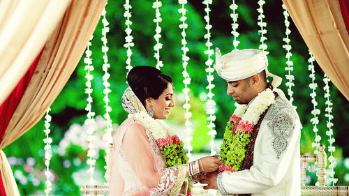 The bride and groom exchanging a special moment during their wedding ceremony beneath floral decor and illuminated hanging garlands | Clarks Amer, Jaipur
