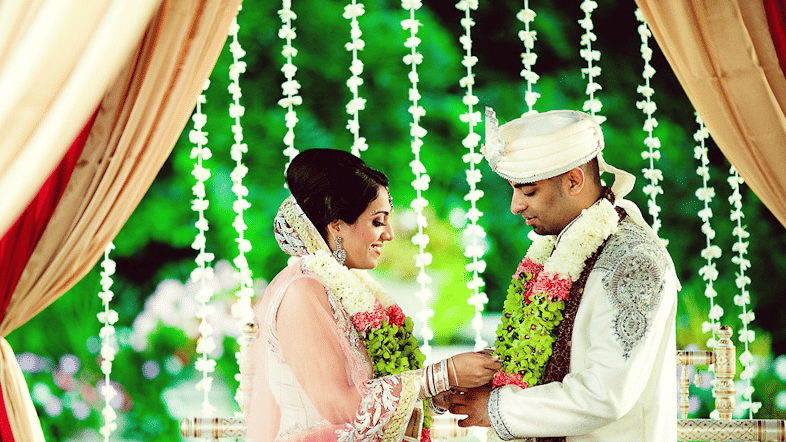 The bride and groom exchanging a special moment during their wedding ceremony beneath floral decor and illuminated hanging garlands | Clarks Amer, Jaipur