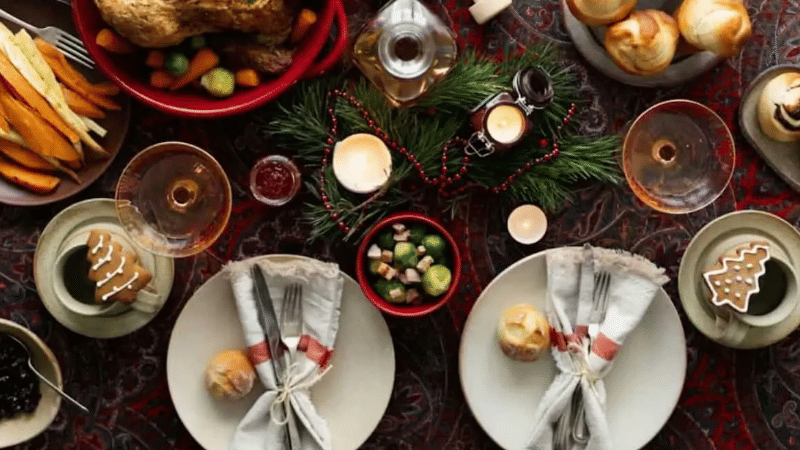 Flat lay of a festive holiday dinner table setting with roast turkey, side dishes, wine glasses, candles, and place settings with Christmas tree gingerbread cookies.