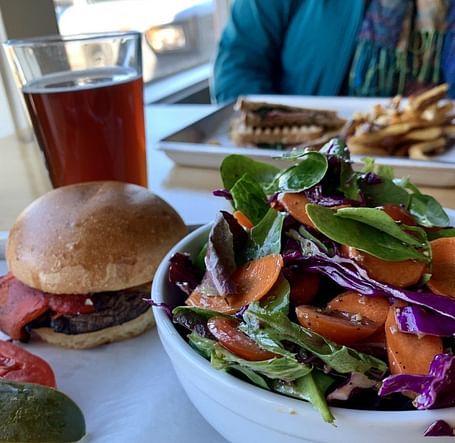 A table with a fresh meal including a burger, a colorful salad, and a glass of beer in a bright dining setting