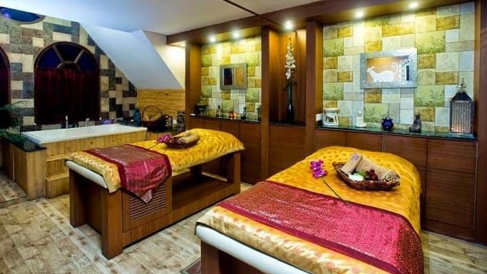 A cosy spa treatment room at Hotel Mount View featuring two massage beds adorned with red and yellow throws. The room features warm lighting, wood accents, and shelves displaying various spa products and decorative items.