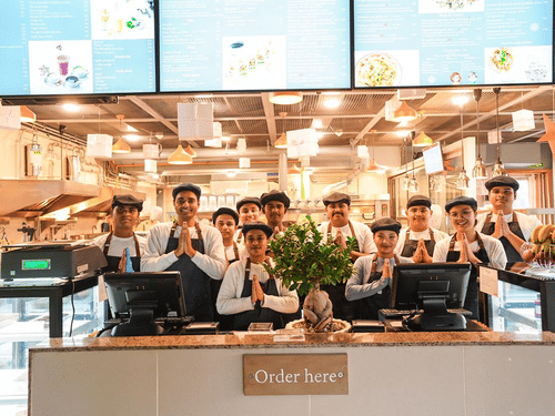 A quick service dining counter at Falaknuma restaurant with digital menu screens, front-facing order counter, staff lined up behind the counter, and kitchen area visible at Clarks Avadh, Lucknow.