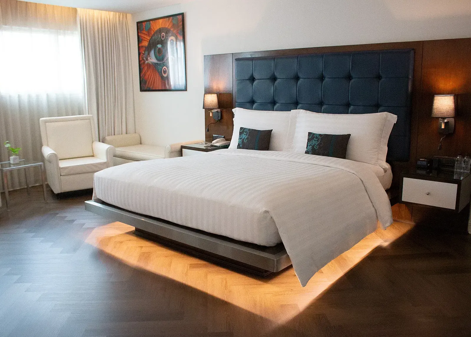 Luxurious hotel suite bedroom with illuminated platform bed, tufted headboard, vibrant artwork, cream armchairs, and warm ambient lighting throughout at Night Hotel Bangkok.